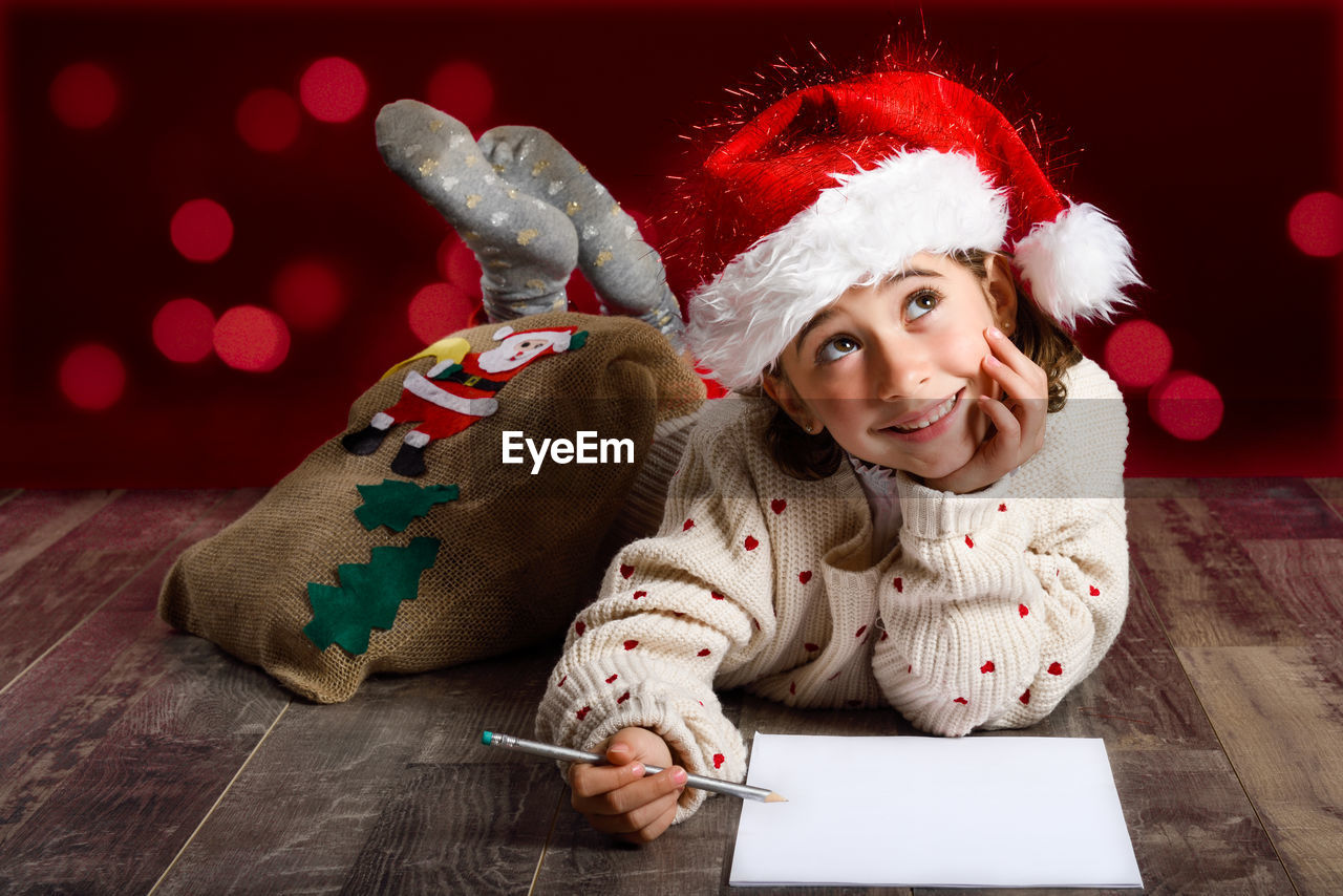 Girl wearing santa hat writing on paper at floorboard against illuminated lights