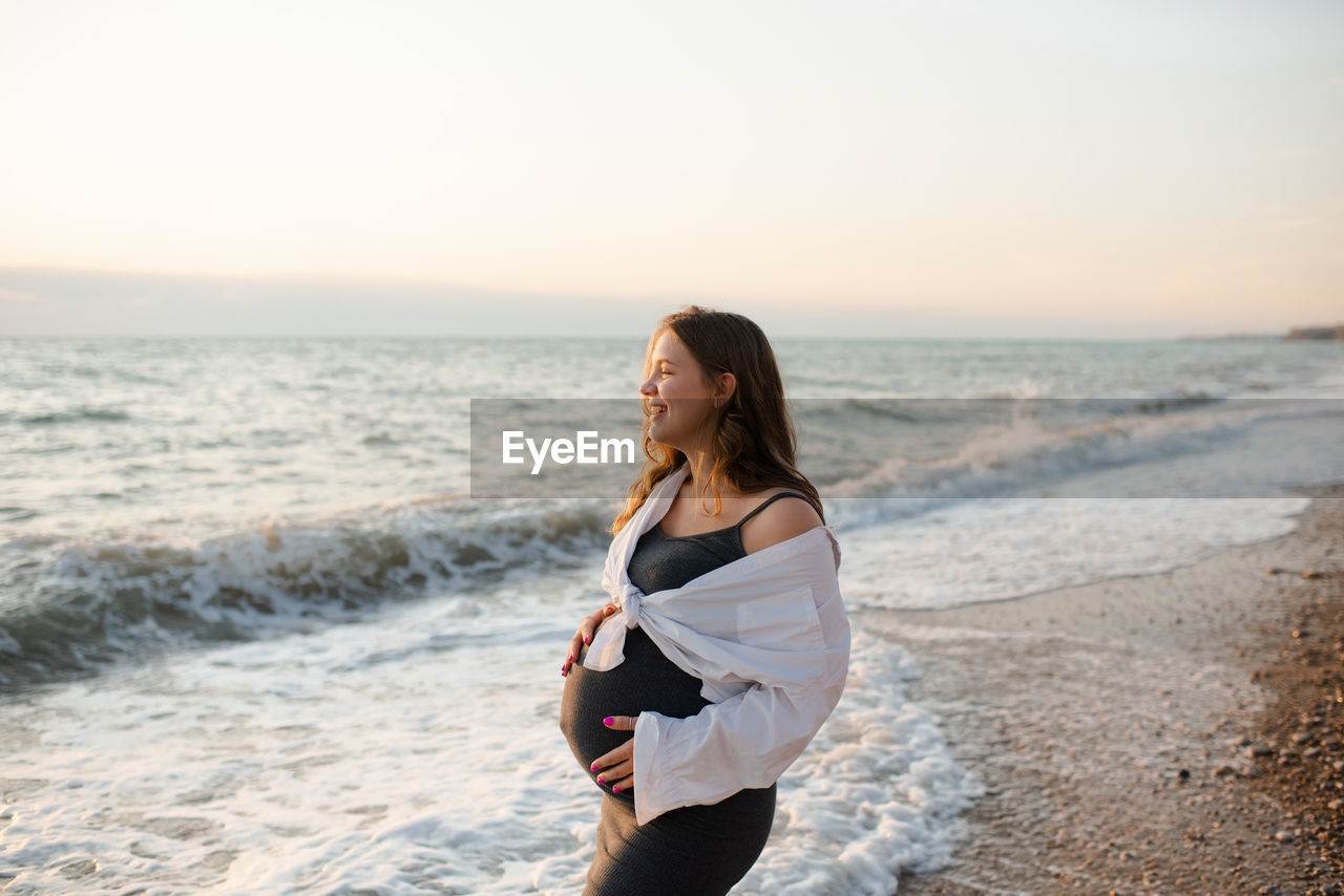 Smiling happy pregnant woman wear casual dress and white shirt standing over sea water outdoor.