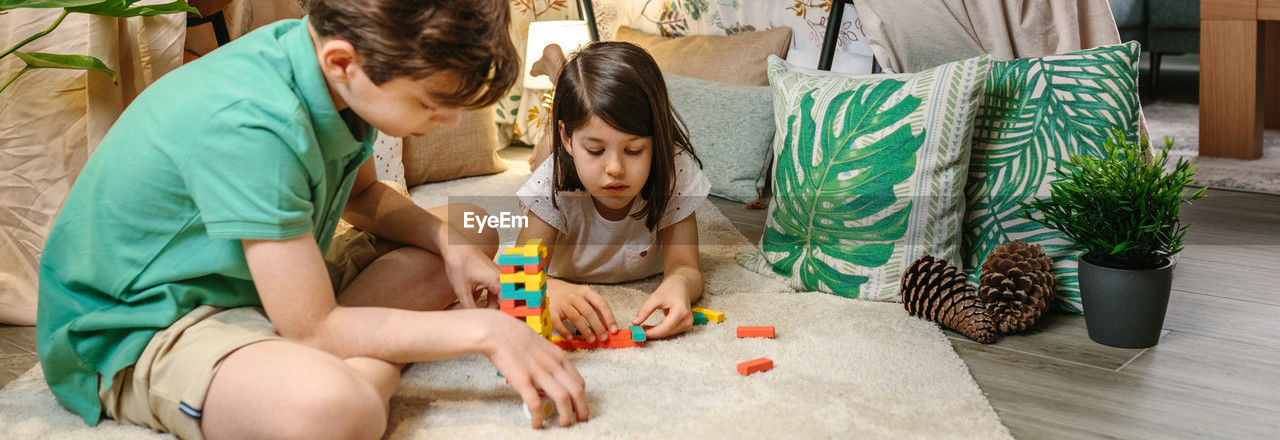 Banner of happy children playing together with stacking piece game in front of homemade tent at home