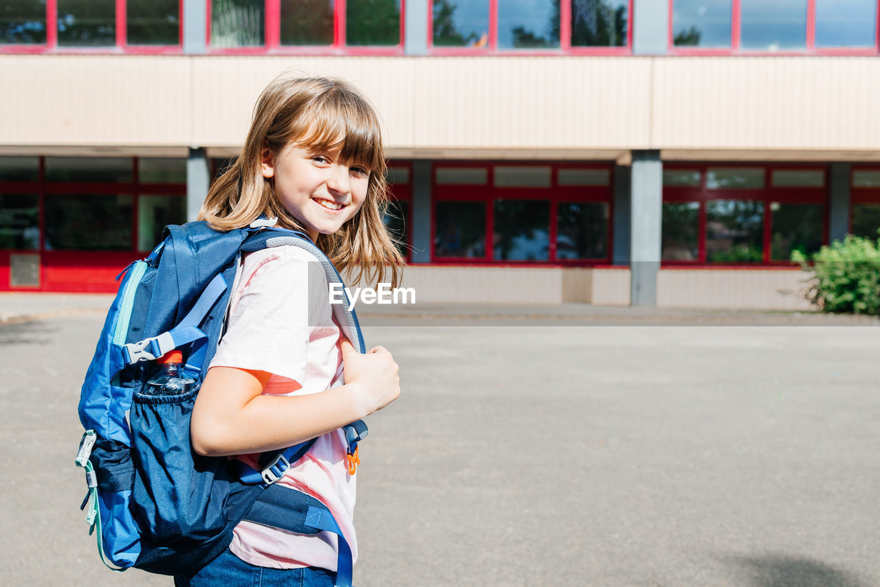Portrait of a happy schoolgirl girl with a backpack on her back against the school.