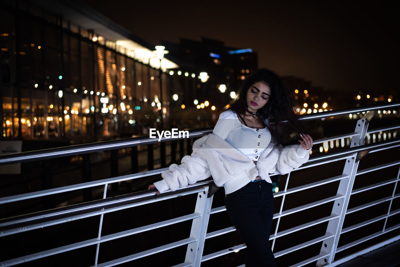 PORTRAIT OF WOMAN STANDING BY RAILING AGAINST ILLUMINATED BUILDING AT NIGHT