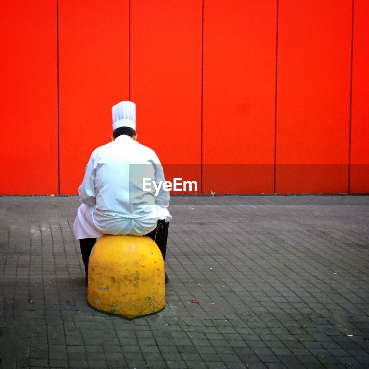 Rear view of chef sitting on yellow bollard against red wall