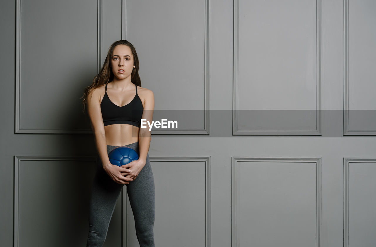 Young woman holding medicine ball standing against wall