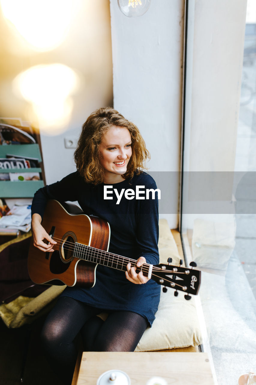 High angle view of smiling young woman playing guitar by window at home