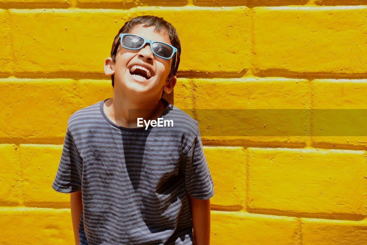 Cheerful boy wearing sunglasses standing against yellow brick wall on sunny day