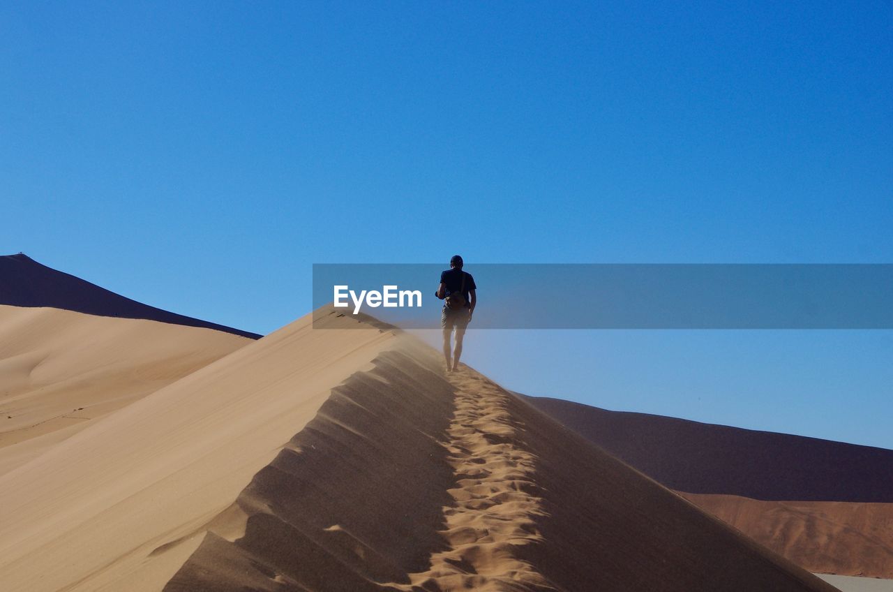 Rear view of man walking in desert against clear blue sky