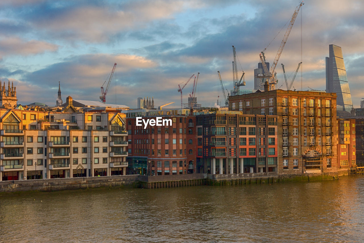 Cityscape by thames river against cloudy sky during sunset