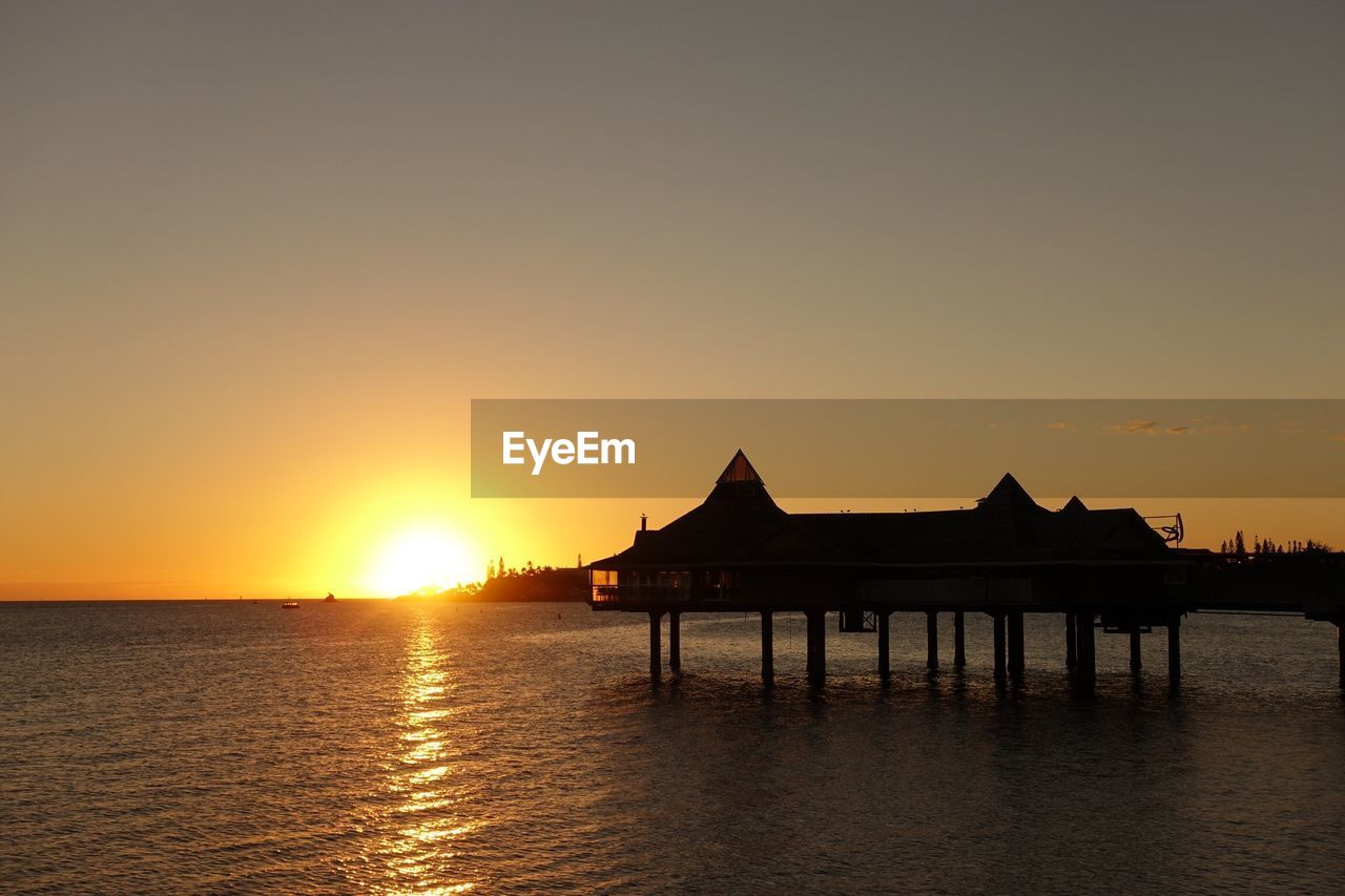 SILHOUETTE BUILT STRUCTURE BY SEA AGAINST ORANGE SKY