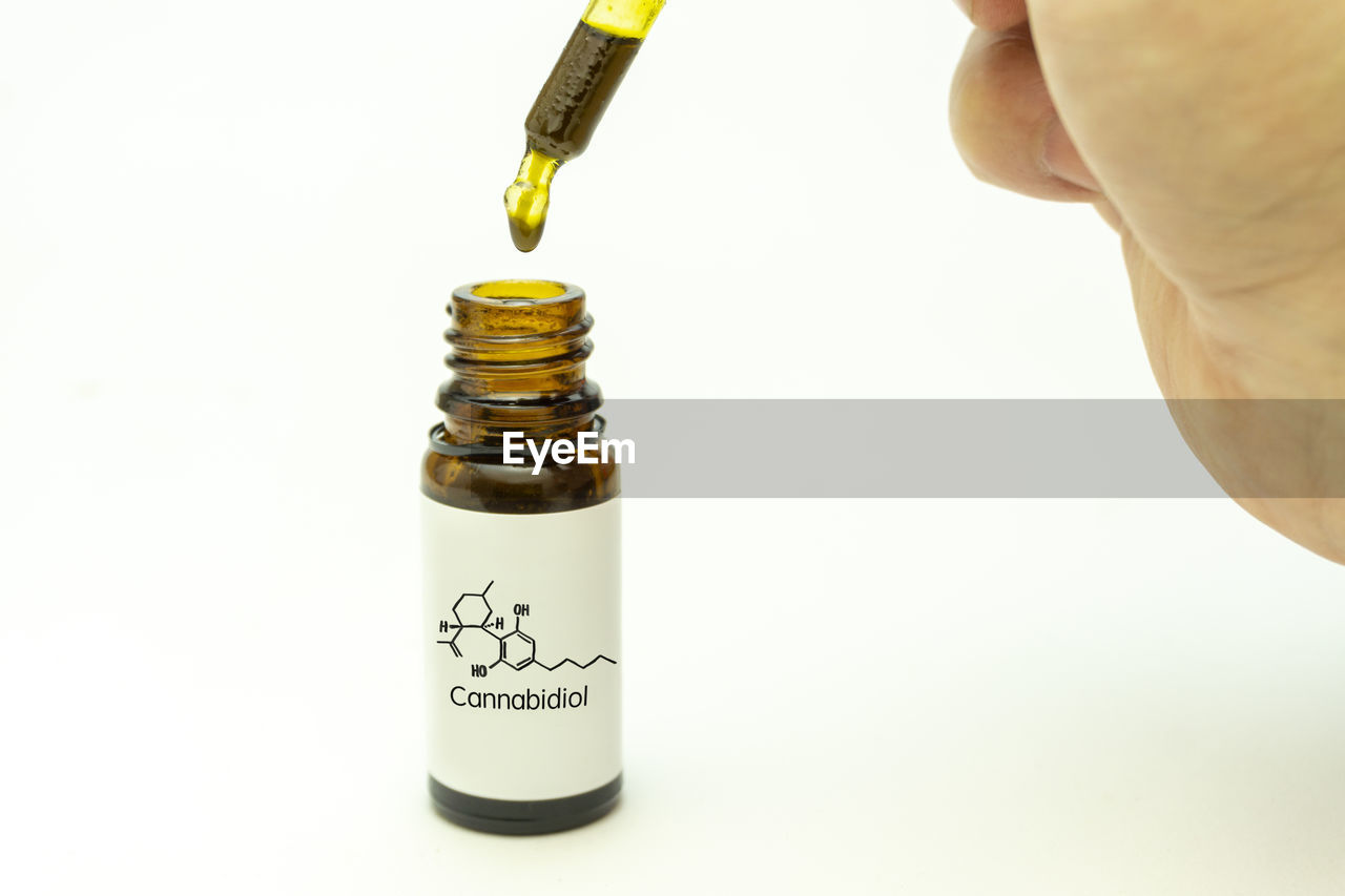 Close-up of hand holding eye dropper over bottle against white background