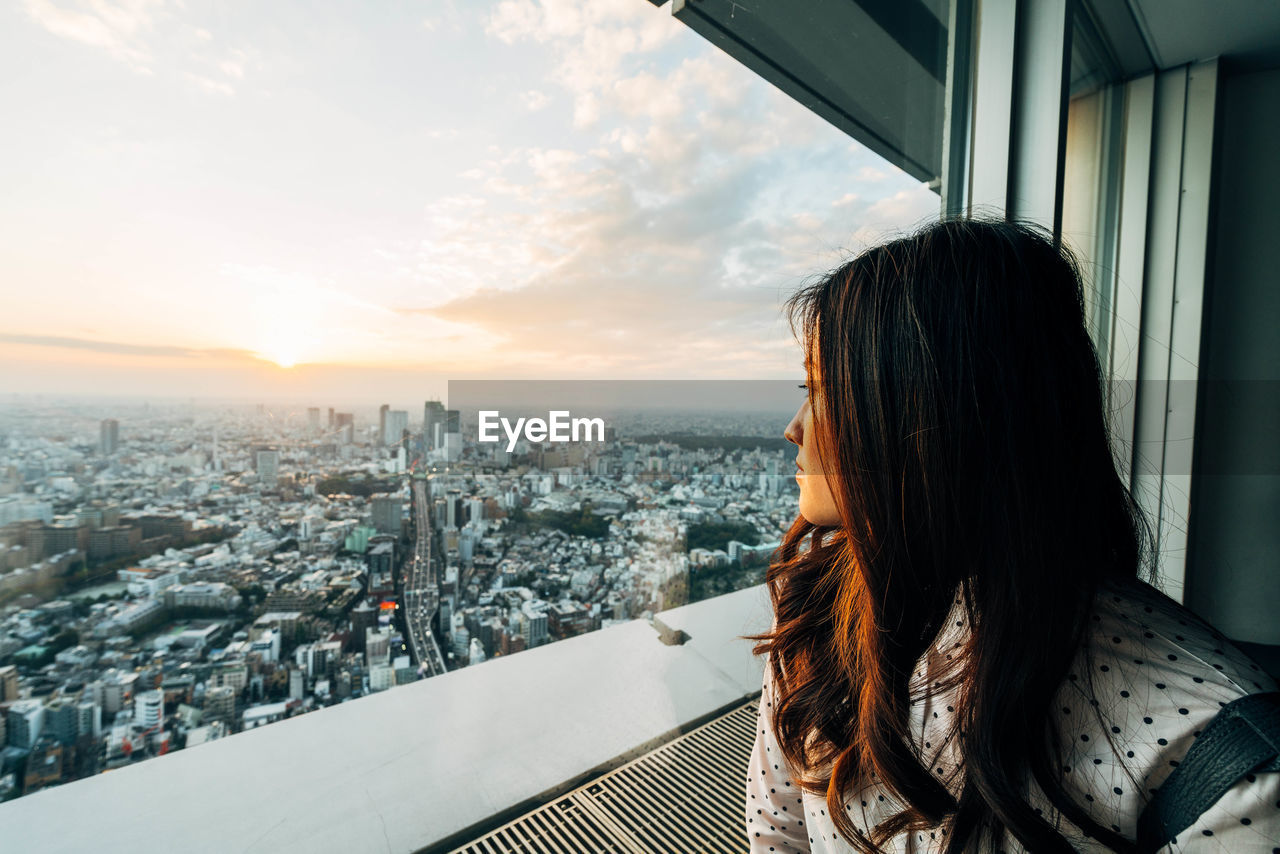 Woman looking at city through window against sky
