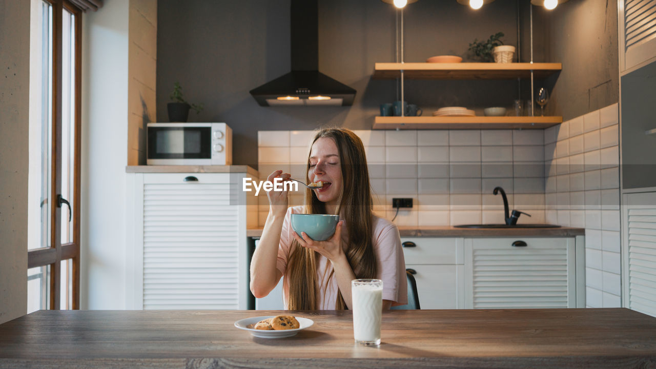 Young female with spoon and bowl enjoying tasty corn rings in kitchen