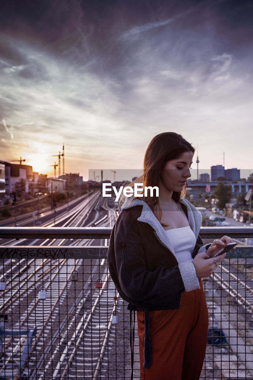 Young woman standing on bridge in city against sky during sunset