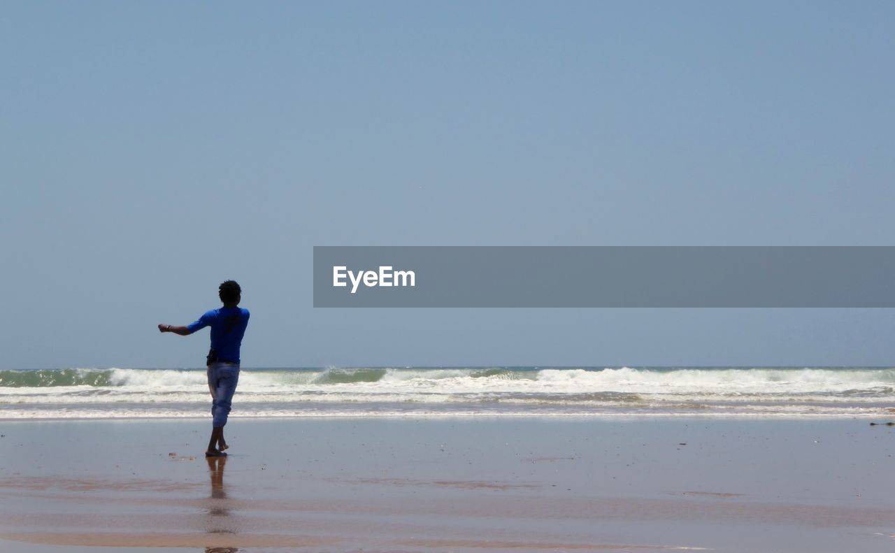 Rear view of man standing at beach against clear blue sky