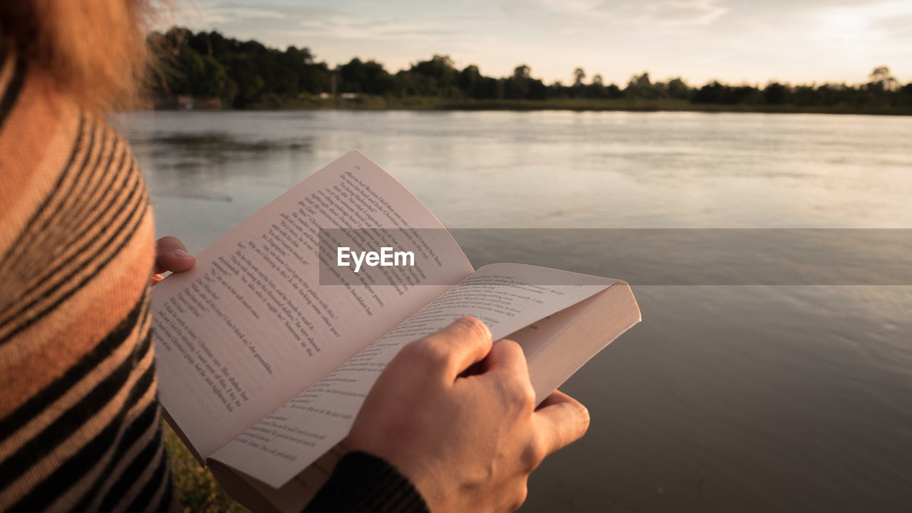 MIDSECTION OF WOMAN HOLDING BOOK BY LAKE