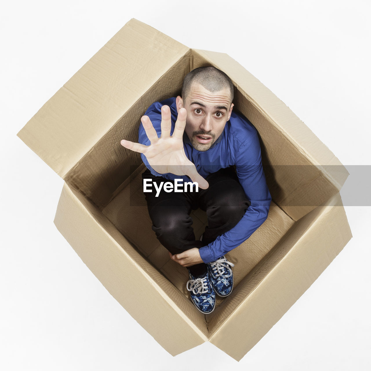 High angle portrait of man gesturing while sitting in cardboard box on white background