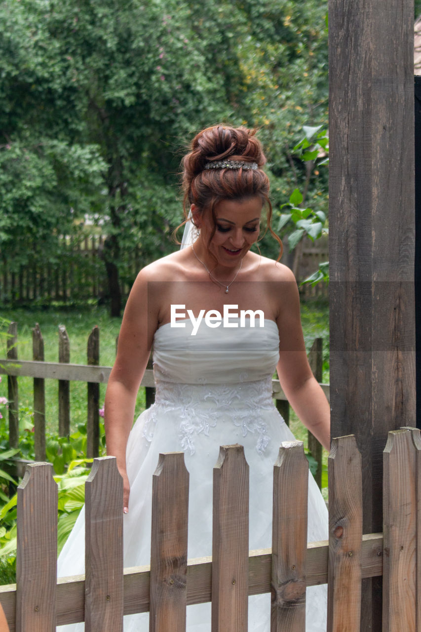 Smiling bride in wedding dress standing by fence against trees