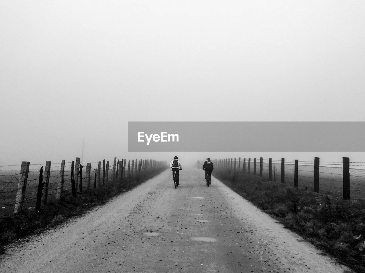 Rear view of people riding bicycle on dirt road against clear sky