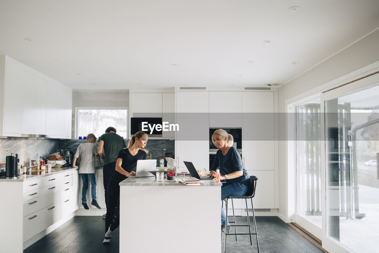 Woman and teenage girl using laptops while man boy standing in kitchen at home