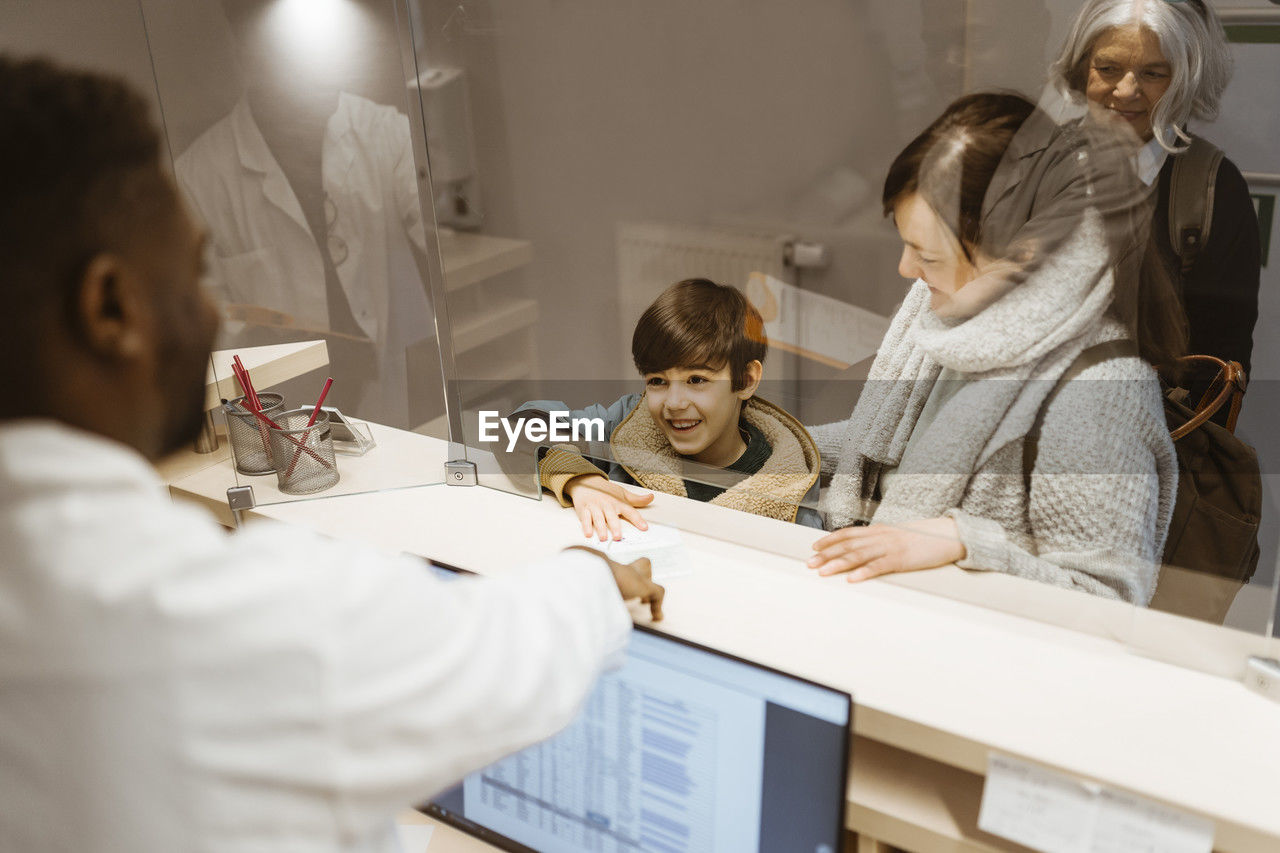 High angle view of smiling boy passing document to male receptionist below transparent shield in clinic