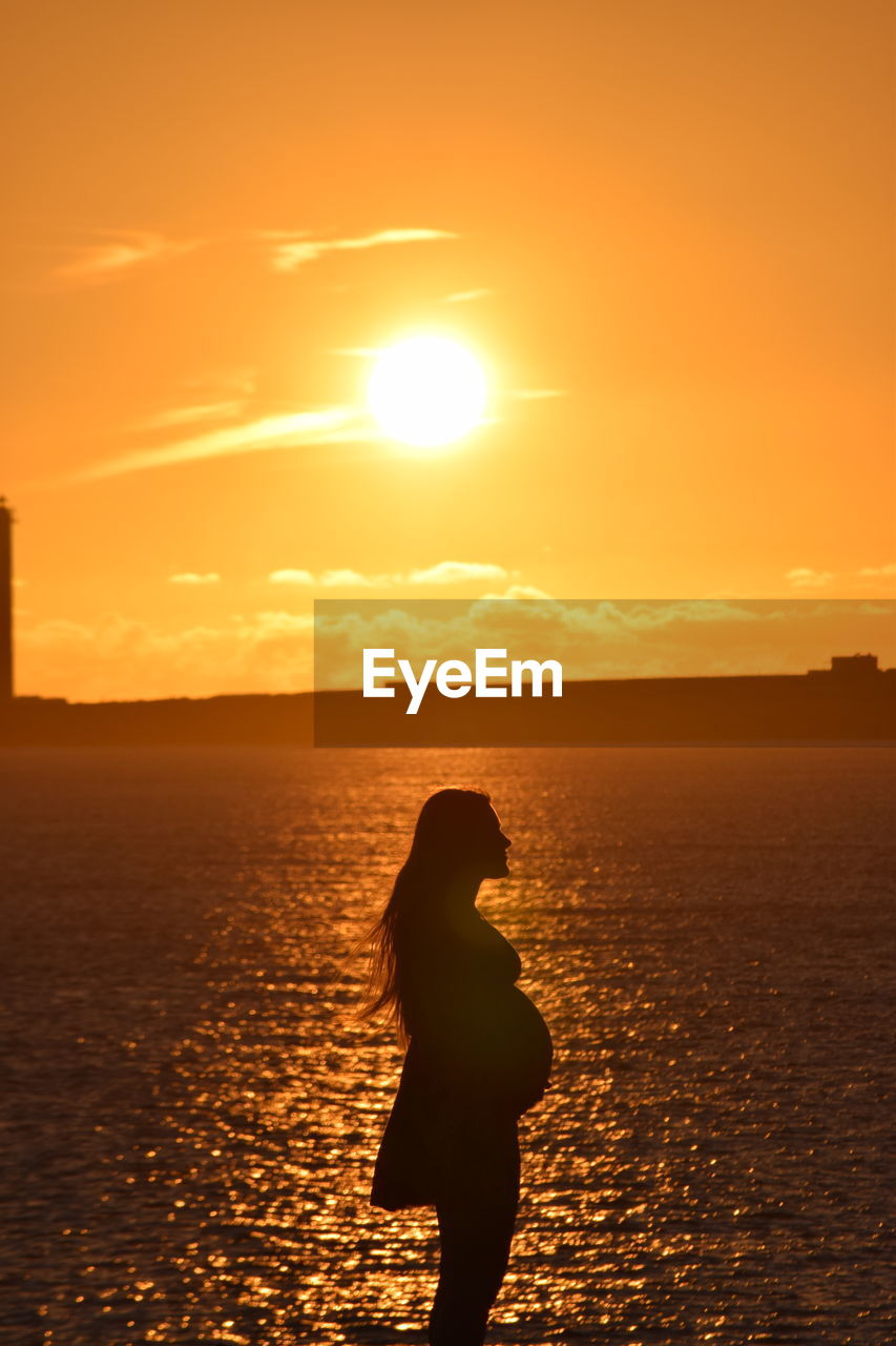 Silhouette pregnant woman standing at beach during sunset