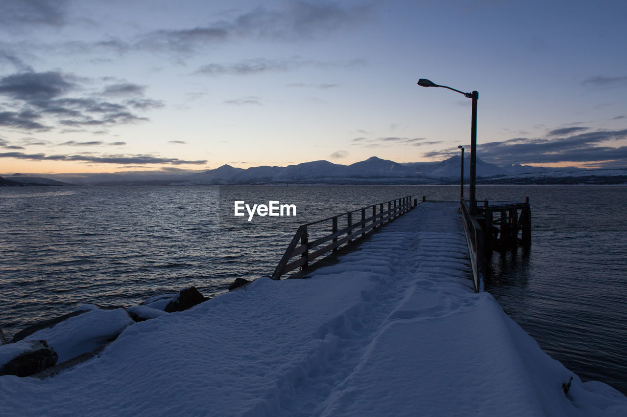 Snow covered pier by lake against sky during sunset