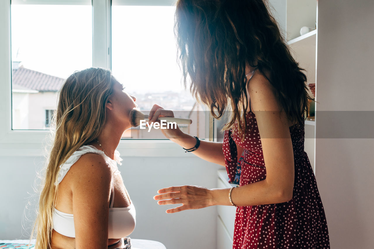 Side view of of delighted female friends with powder and brush doing makeup at home and looking at each other while laughing