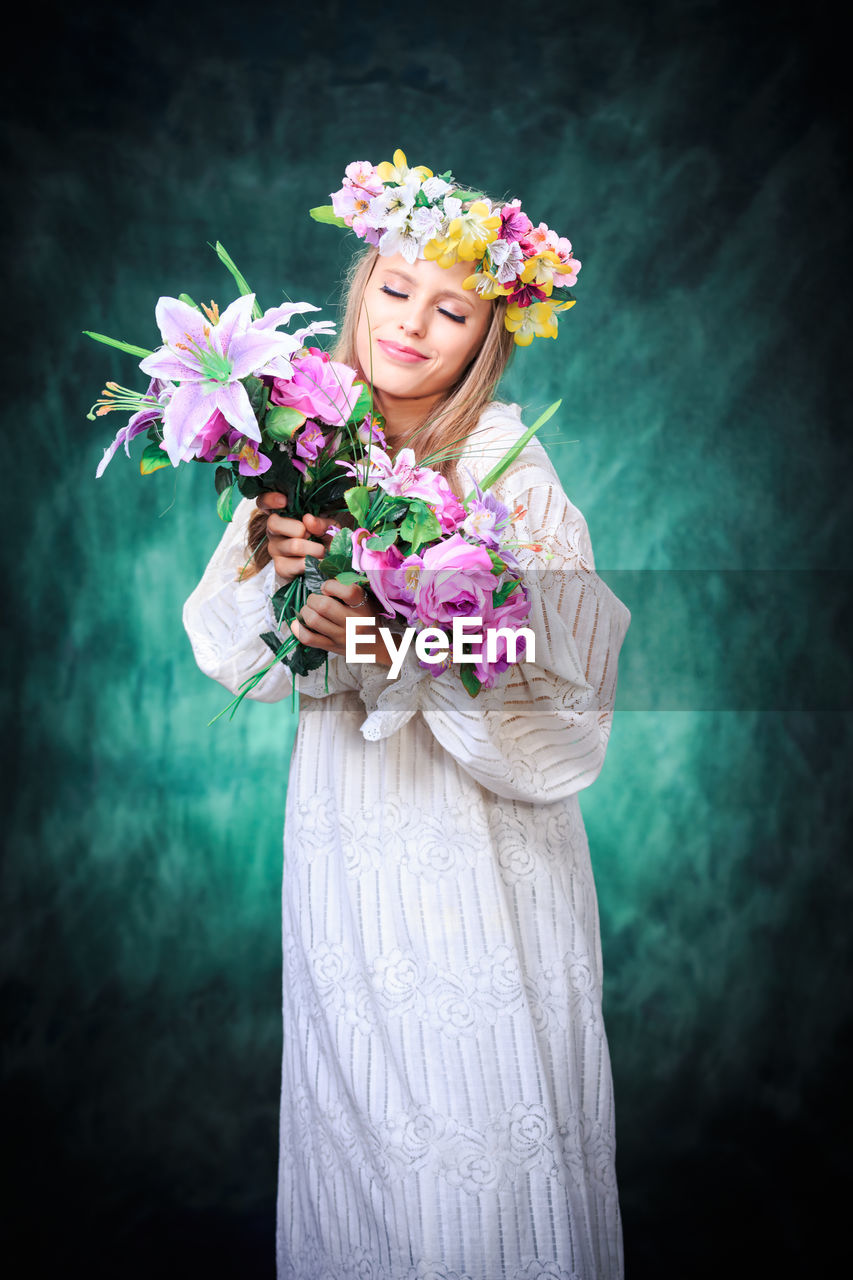 Smiling teenage girl with eyes closed wearing dress and tiara holding bouquet against wall