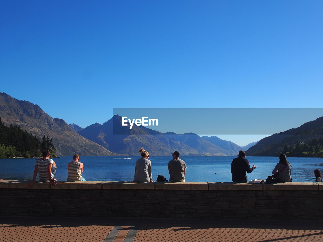 Rear view of people sitting on retaining wall by sea against clear blue sky