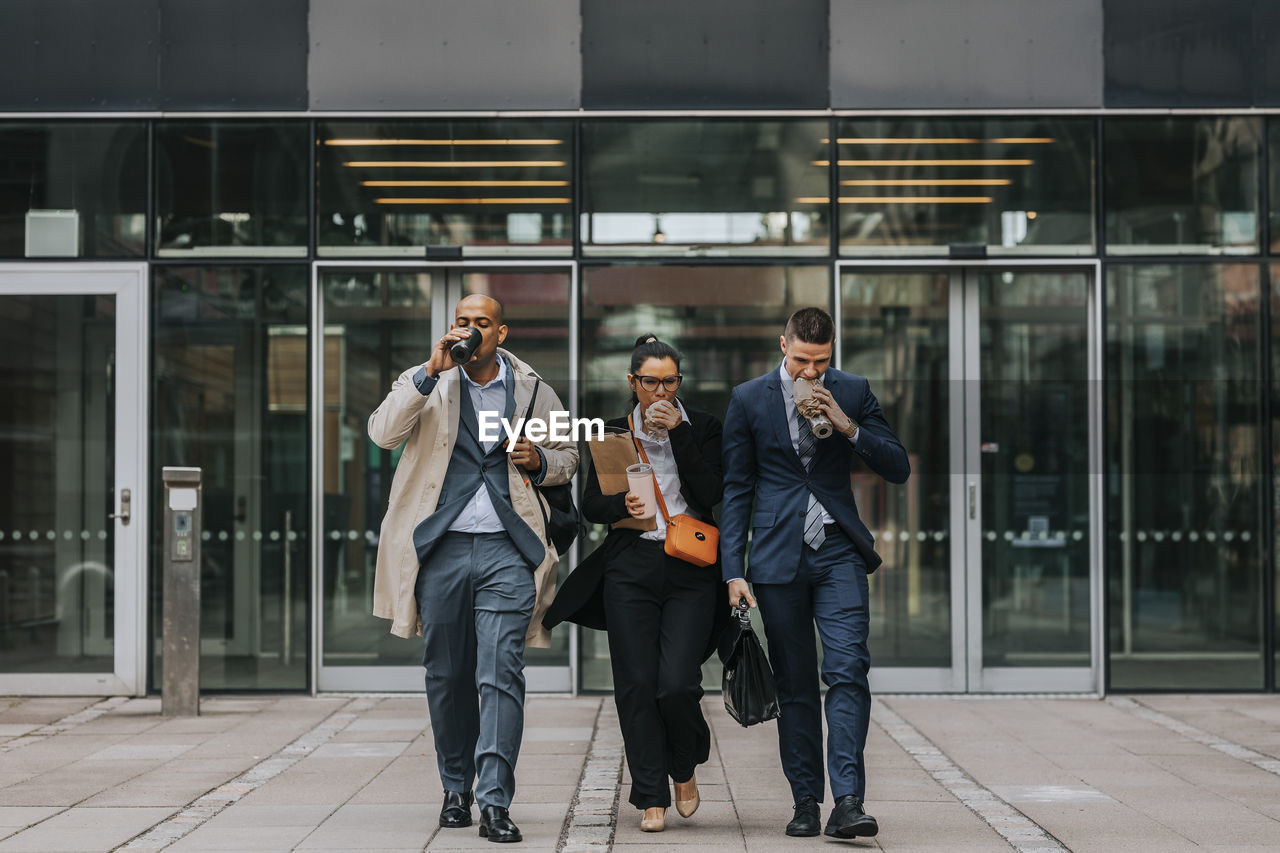 Full length of male and female business colleagues having food while walking in front of office building