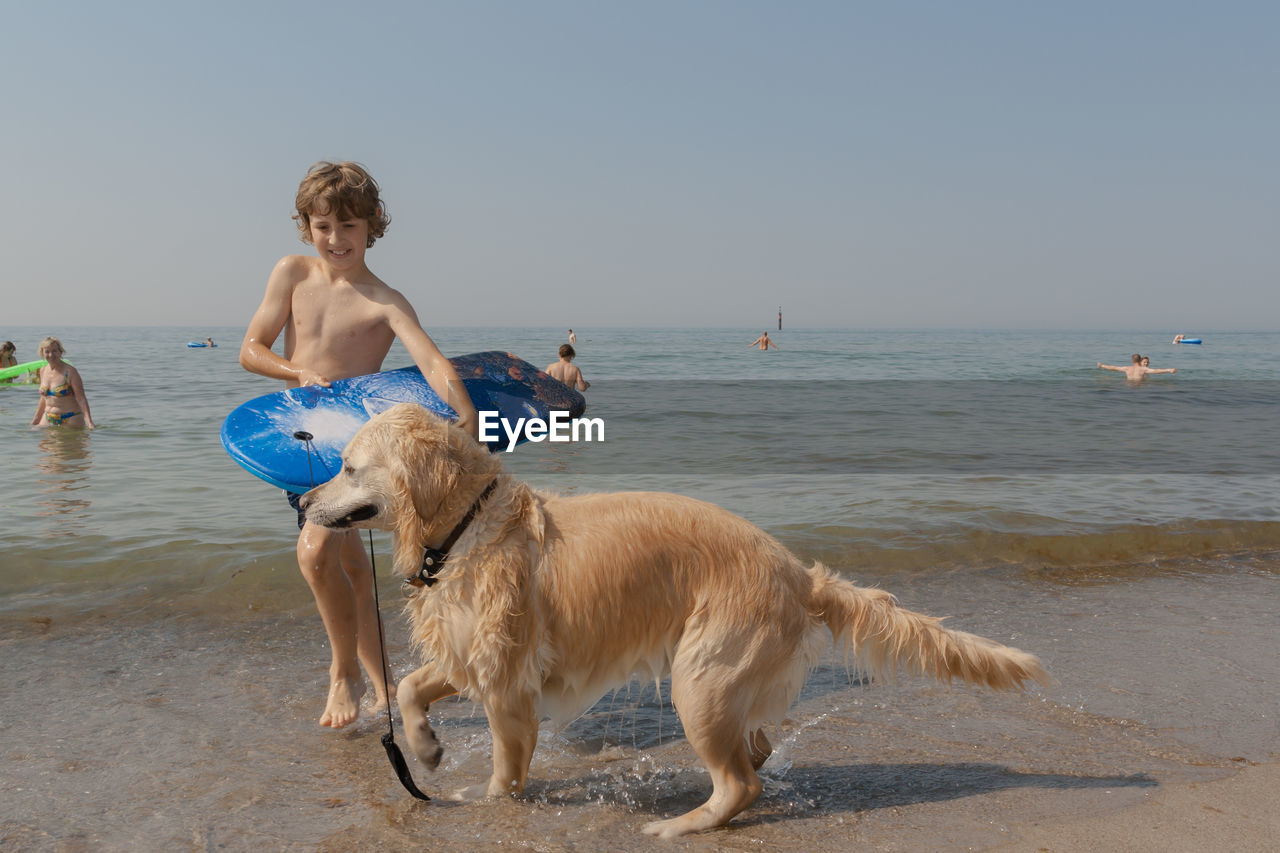 Shirtless boy carrying surfboard walking with golden retriever walking at beach against sky