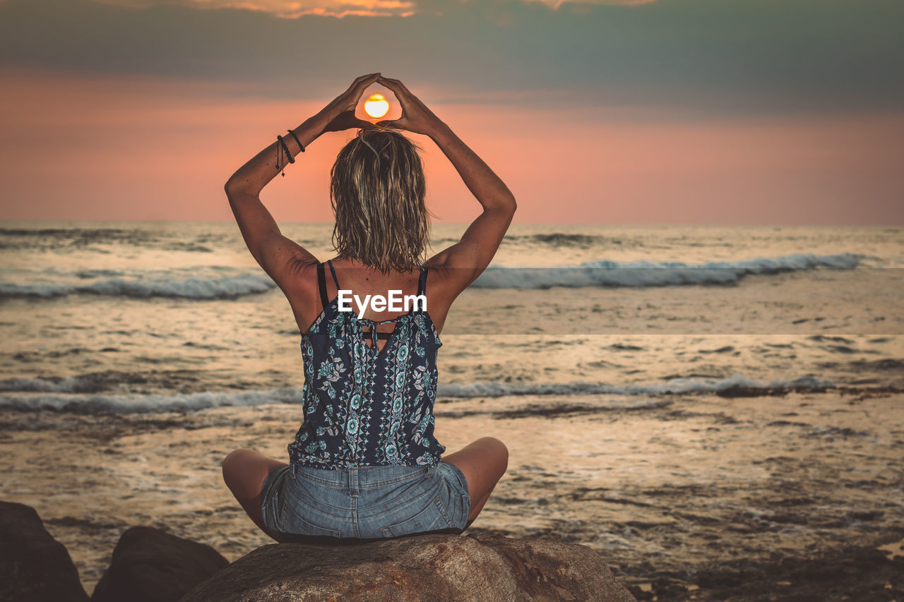 Woman doing yoga on rock at beach against sky during sunset