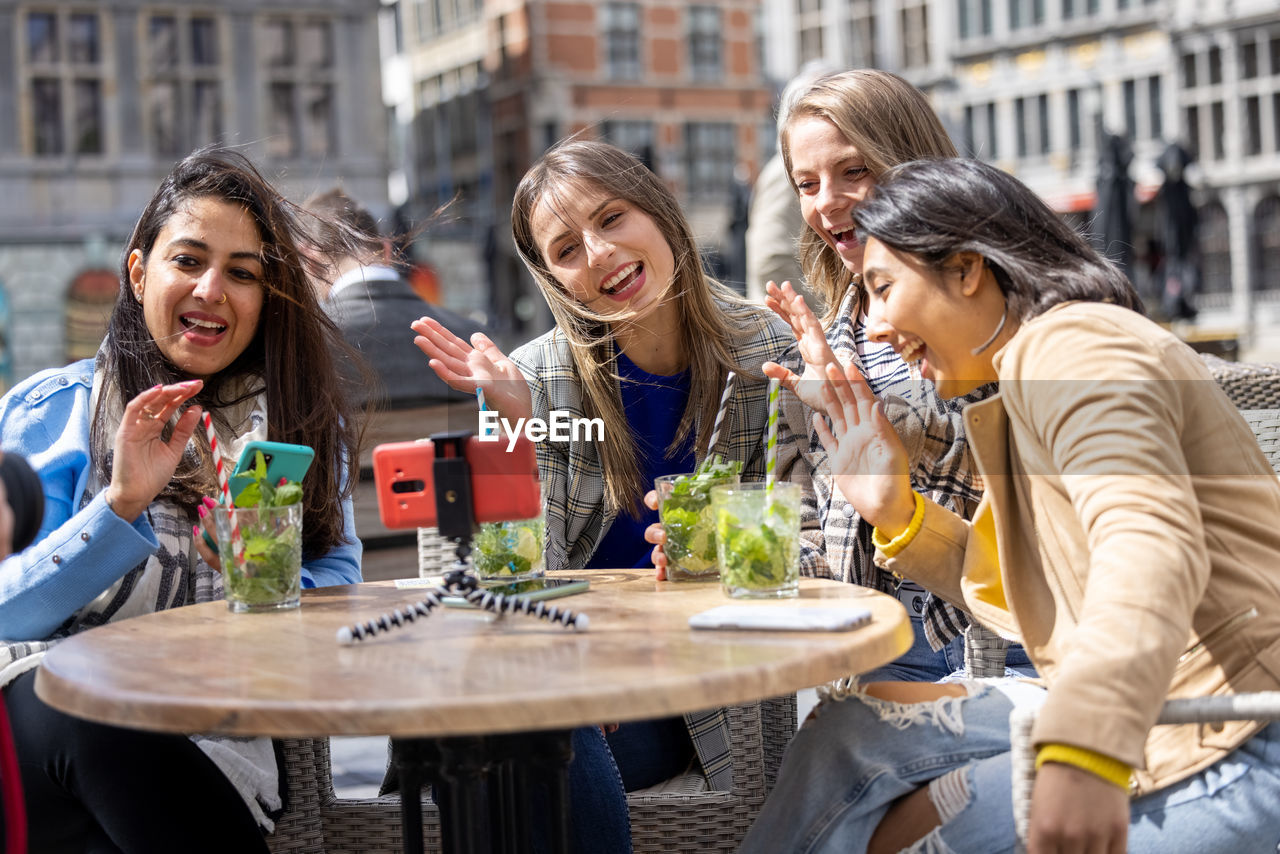 Four multi ethnic female friends sitting at a cafe terrace using mobile phone for a video call