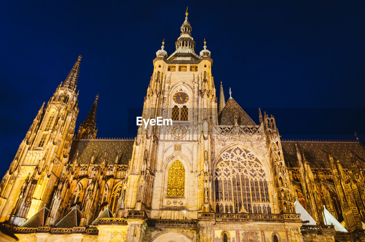 Dramatic view of the prague castle illuminated by warm yellow lights in a dark blue sky.