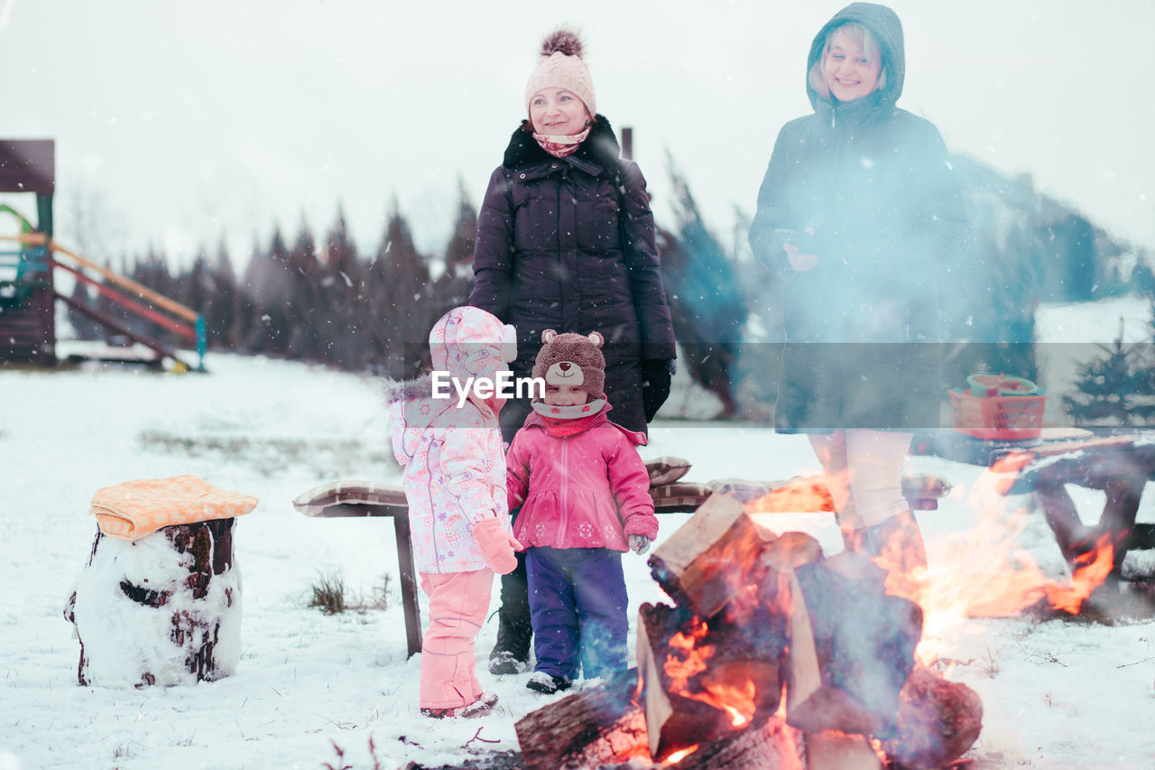 Mother with children standing on snow covered field at campfire against sky
