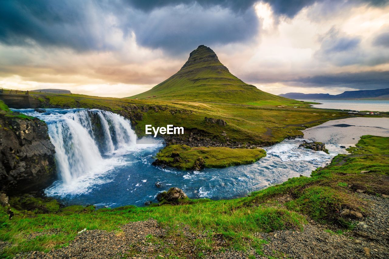 Scenic view of waterfall by kirkjufell against cloudy sky during sunset