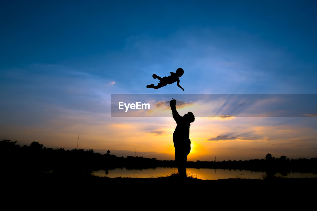 Silhouette father throwing son into air while standing at lakeshore against sky during sunset