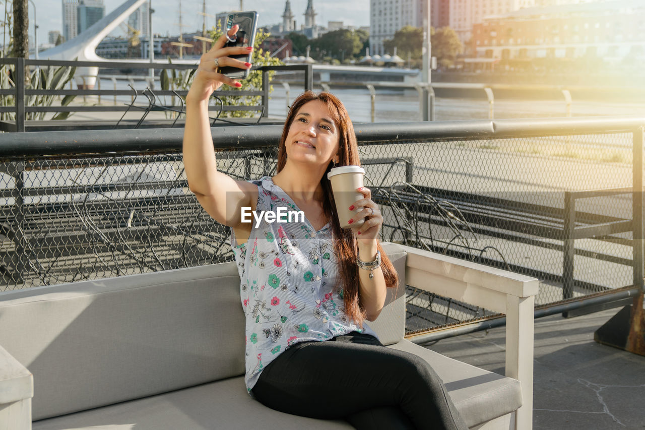 Hispanic woman in coffee shop taking a selfie with her phone holding cup of coffee sitting.