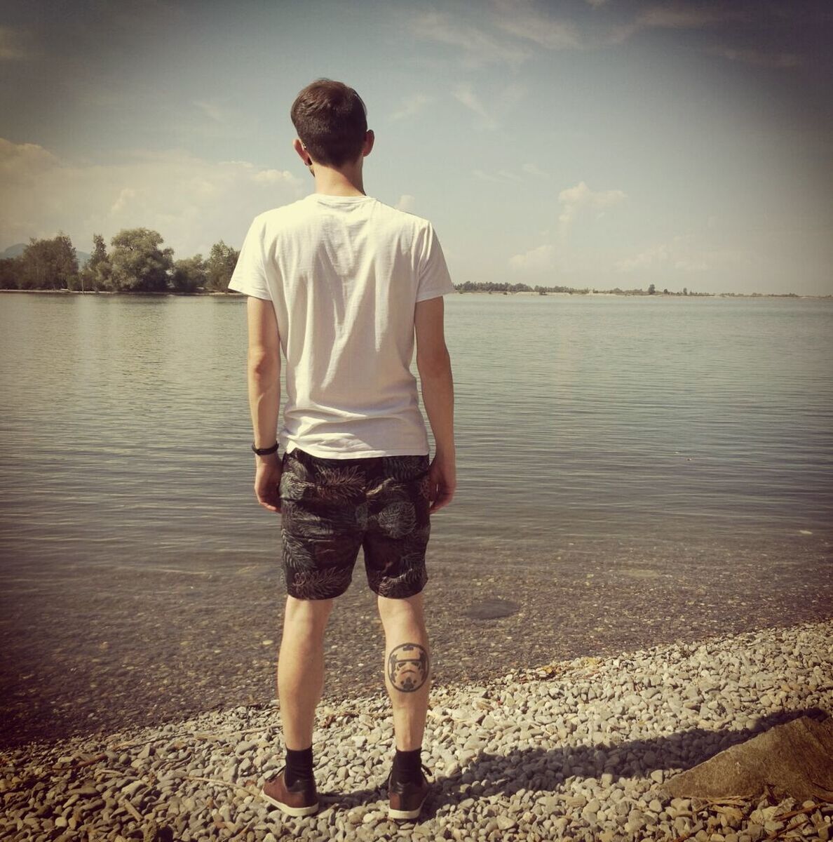 Rear view of man with tattoo on leg standing at shore against sky
