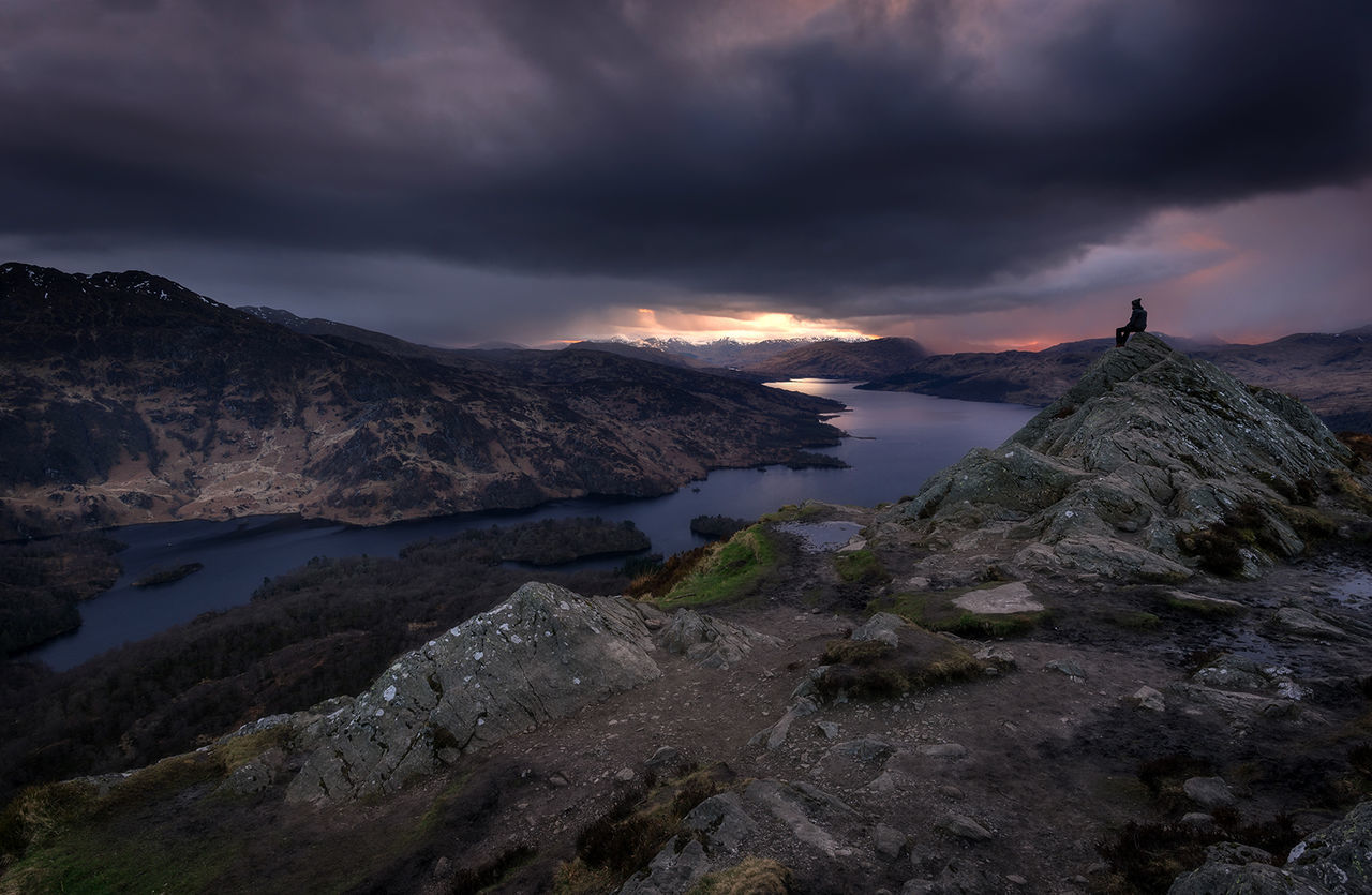 Hiker sitting on rock formation by lake against cloudy sky at dusk