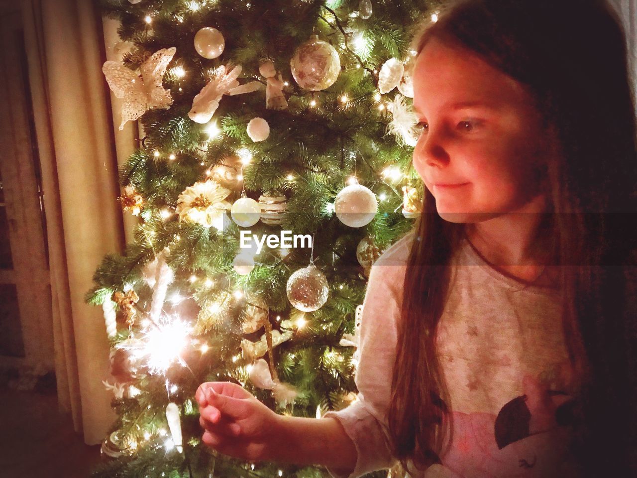 Smiling girl holding burning sparkler while standing by illuminated christmas at home