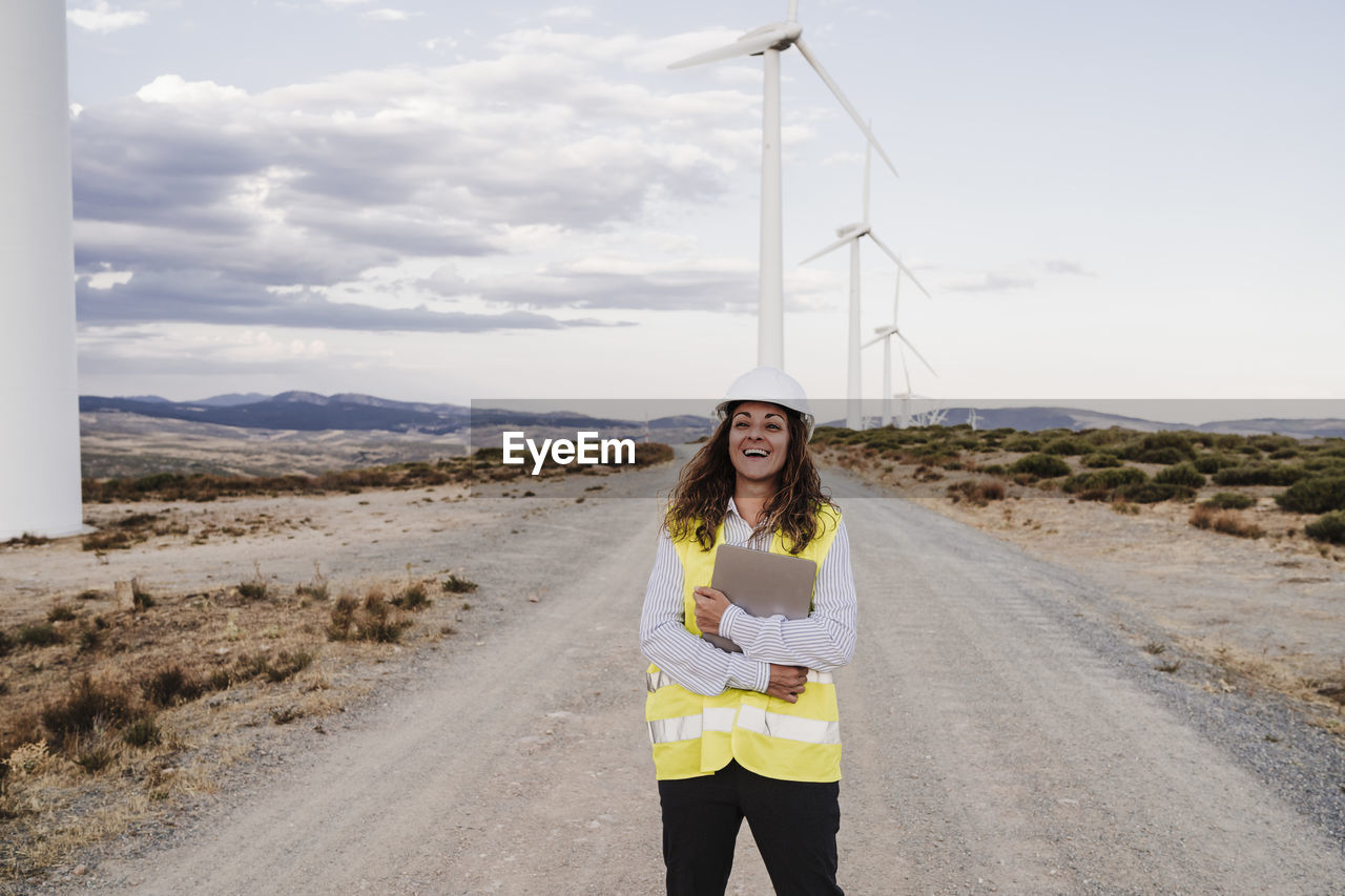 Happy engineer holding laptop standing on dirt road at wind farm