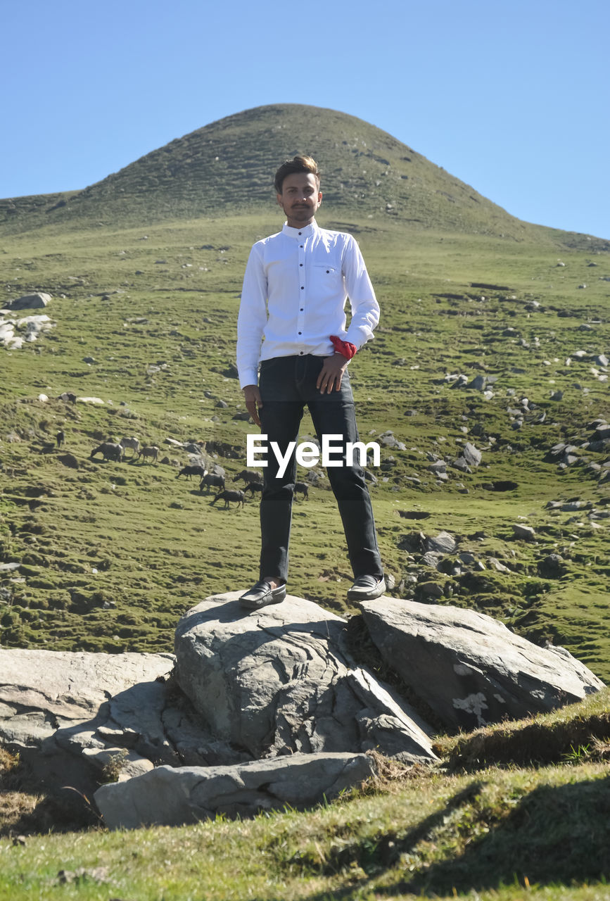 A handsome indian young guy looking at camera while standing over rock in the mountain