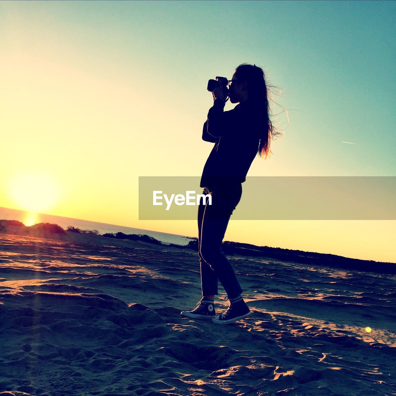 Side view of silhouette woman photographing at beach against clear sky during sunset