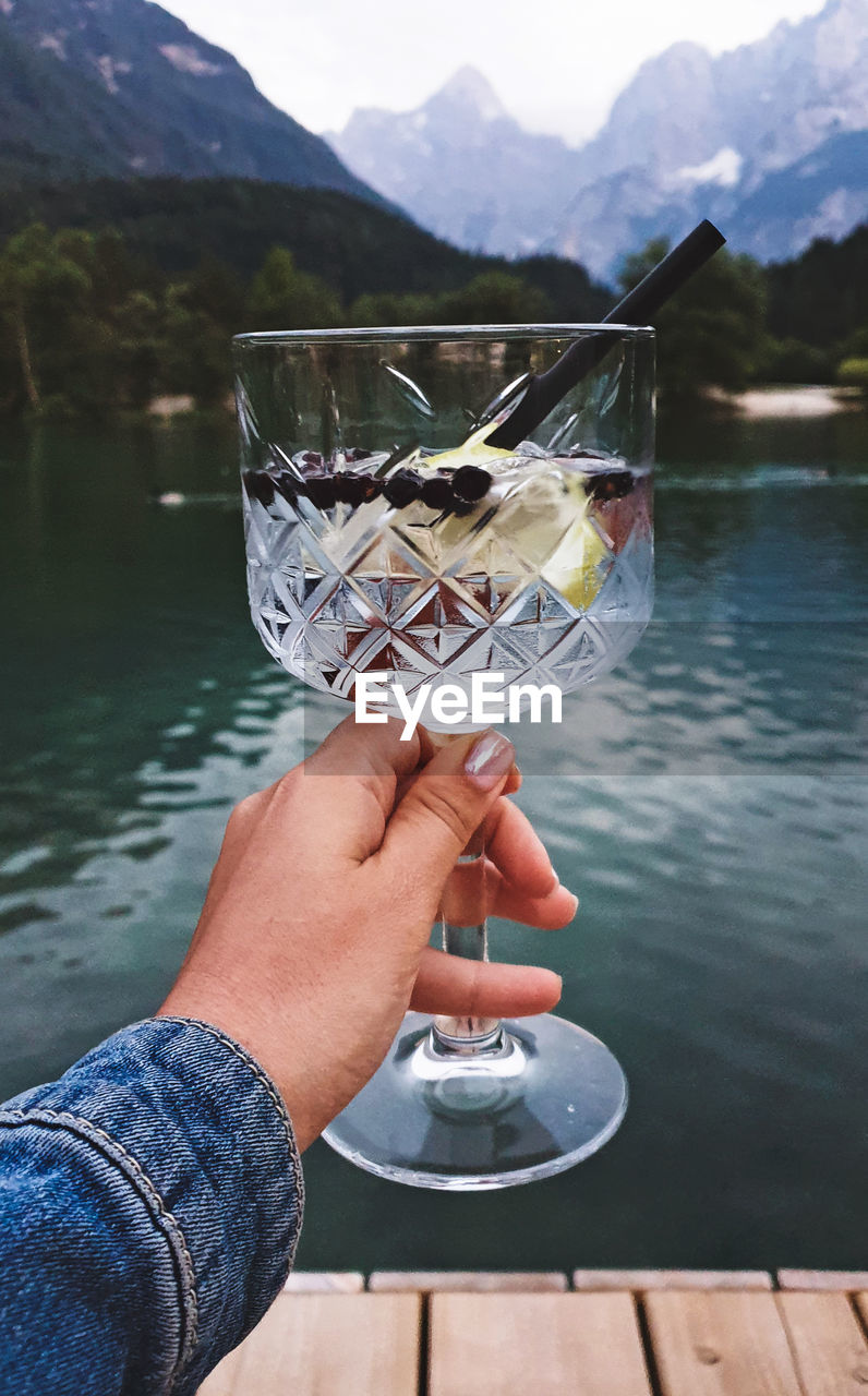 Personal perspective of woman holding glass with drink. lake, mountains in background.