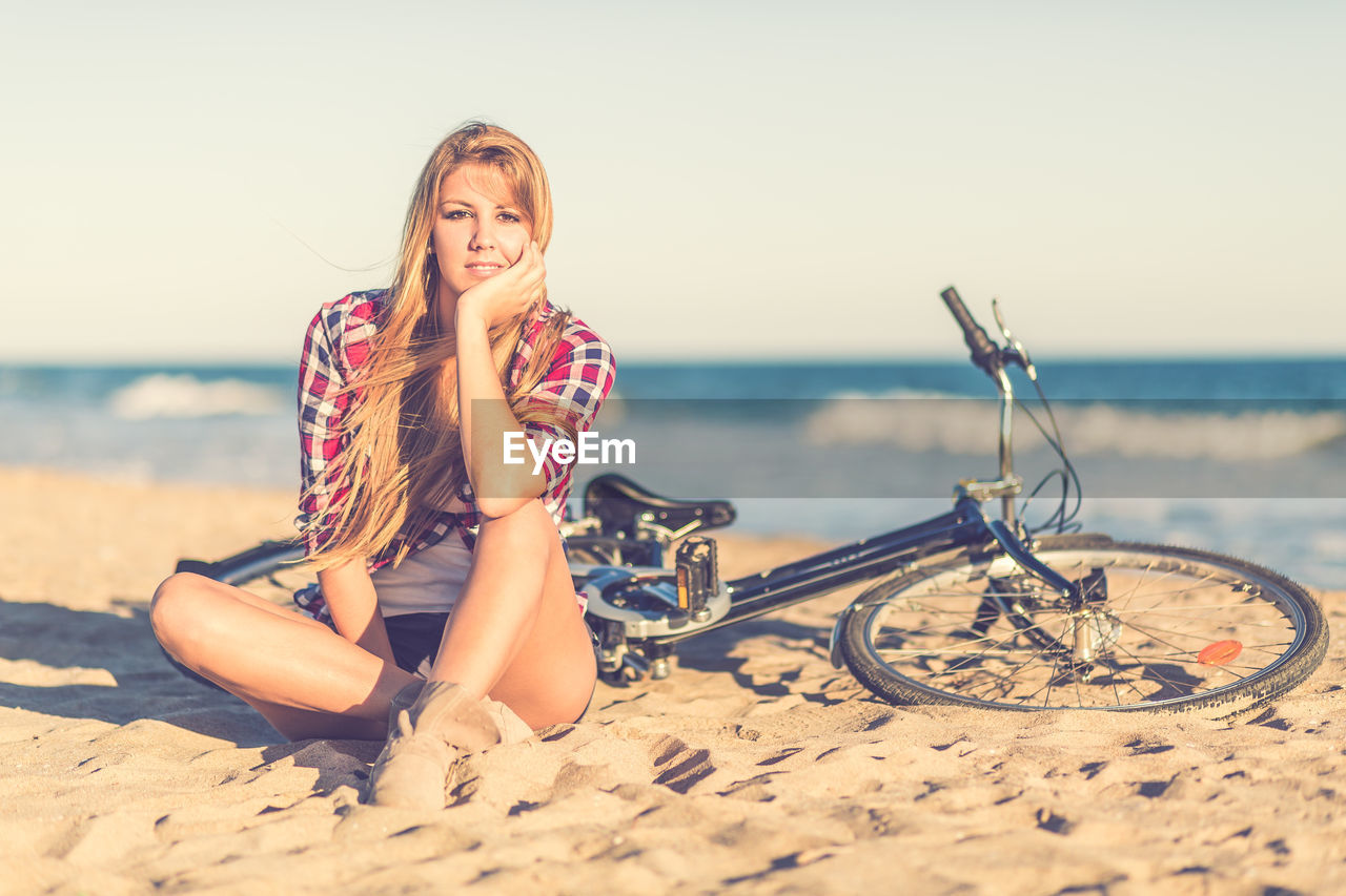 Portrait of woman with bicycle sitting at beach against sky