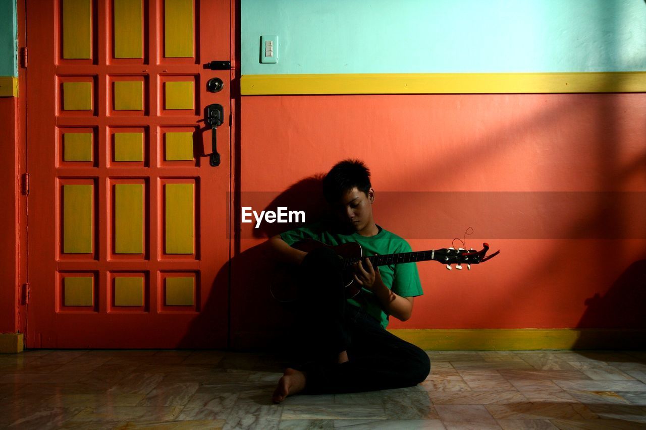 Young man playing guitar while sitting against wall