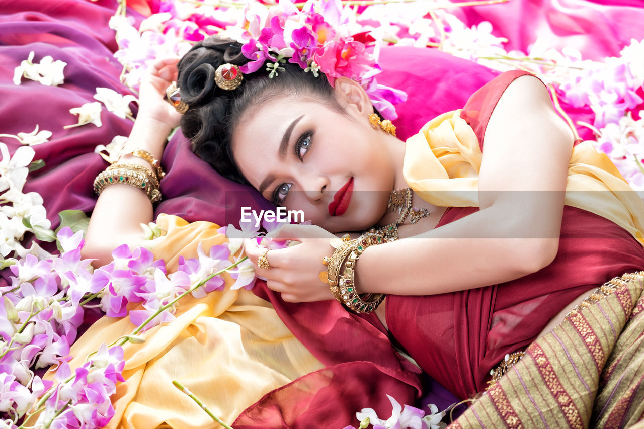 High angle view of beautiful woman wearing traditional clothing and jewelry while lying with flowers on bed
