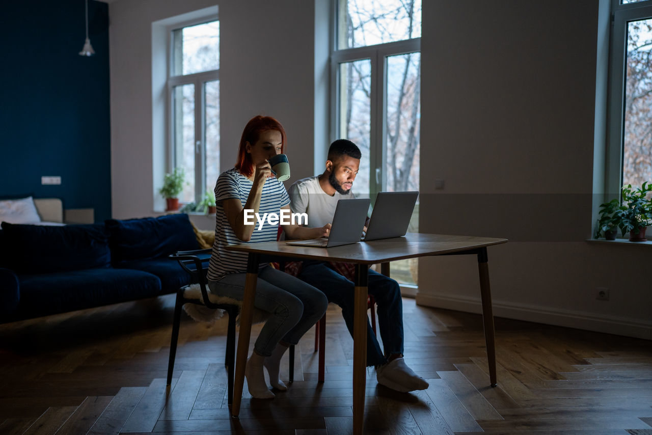 Young modern freelancing couple sitting at table in living room working from home together
