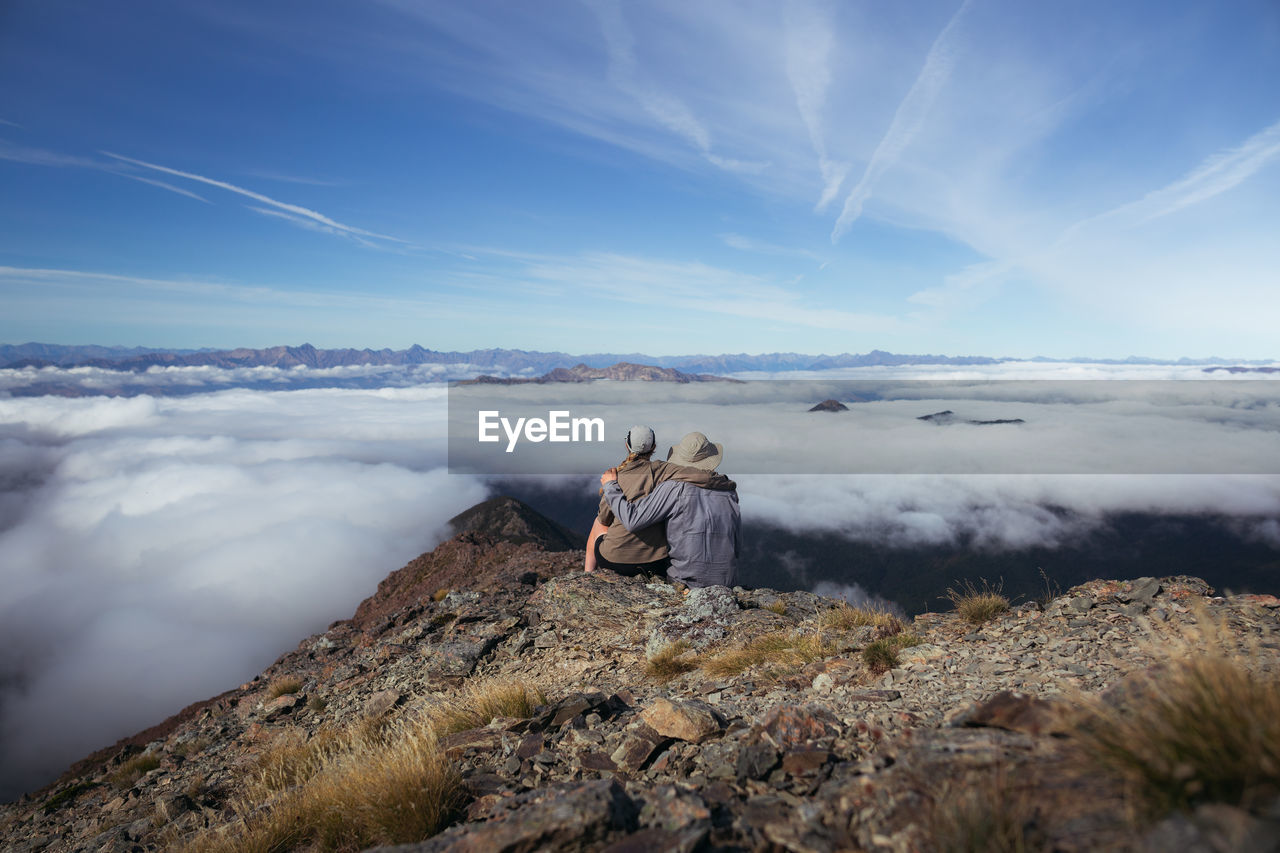 Rear view of man and woman sitting on rock against sky