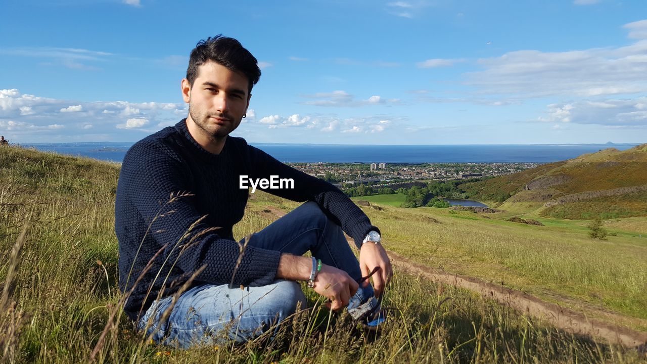 Portrait of young man sitting on field against sky